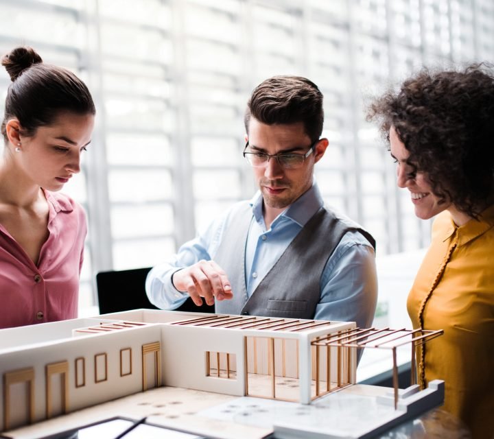 A group of young architects with model of a house standing in office, working and talking.